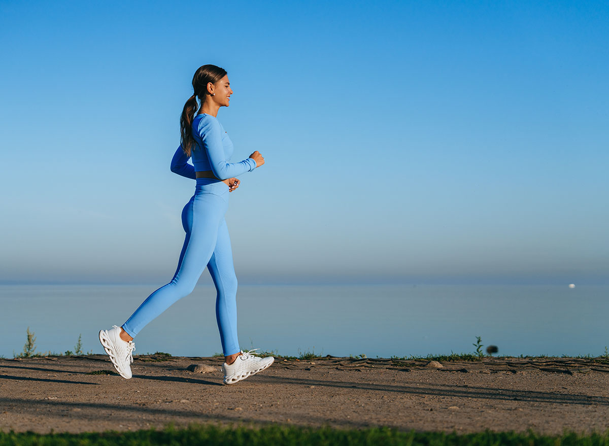 A woman in a vibrant blue outfit briskly walks along a serene waterside path, basking in the calmness of a clear morning sky.
